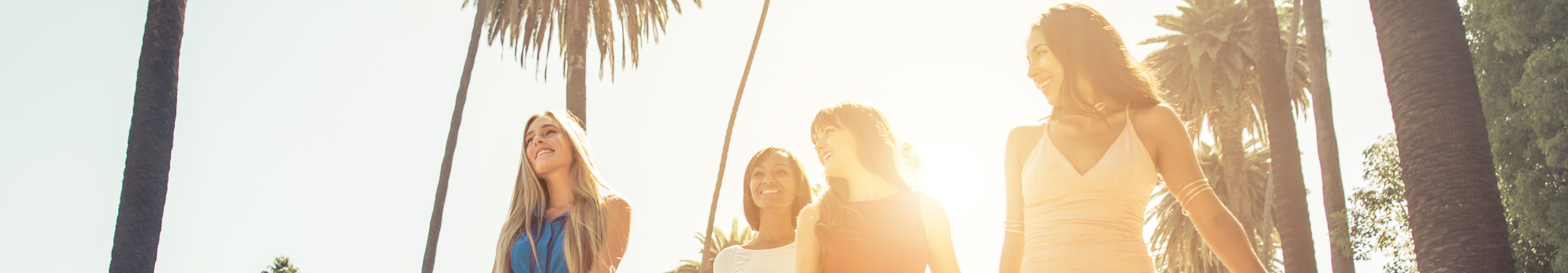 Vier junge Frauen in bunten Sommerkleidern gehen lachend mit Einkaufstüten eine von Palmen gesäumte Straße entlang, während die Sonne im Hintergrund hell scheint.