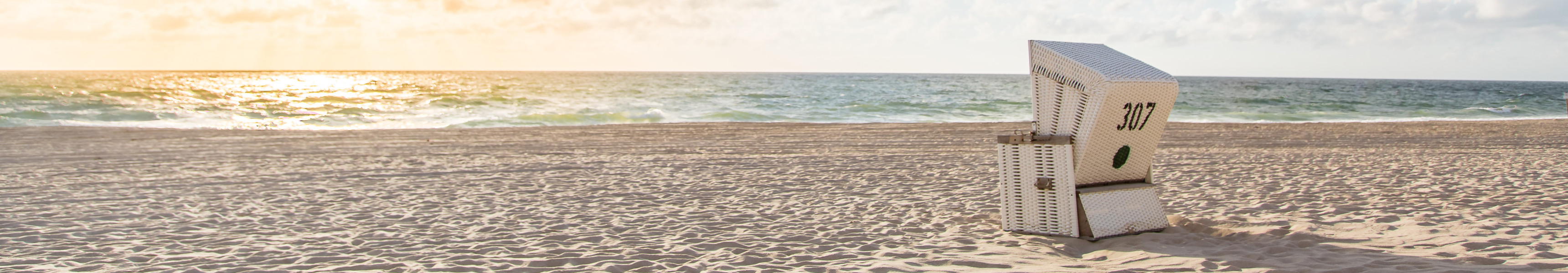 Ein einzelner Strandkorb mit der Nummer 307 steht am leeren Sandstrand von F&ouml;hr, w&auml;hrend die Sonne hinter Wolken am Horizont &uuml;ber der Nordsee untergeht.